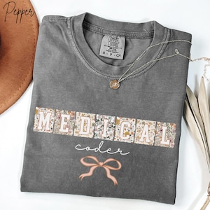 May include: A gray t-shirt with the words "MEDICAL coder" in a floral design. The shirt has a small pink bow graphic and a silver necklace with a pendant. The shirt is a Comfort Colors brand.