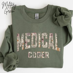 May include: Military green sweatshirt with the words "MEDICAL CODER" in a floral design. The sweatshirt is a solid color with a crew neck and long sleeves. The text "Military Green" is in a script font in the upper left corner.