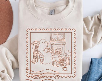 May include: A sand-coloured sweatshirt featuring a brown illustration of a ghost reading in a cosy setting. The design includes a fireplace, pumpkins, a cat, and a cup of coffee, within a postage stamp frame.