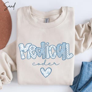 May include: A sand-colored sweatshirt with the words "Medical coder" in blue, patterned lettering. The word "Medical" has a mix of patterns, including floral, leopard print, and checkered designs. A blue heart graphic is below the text.