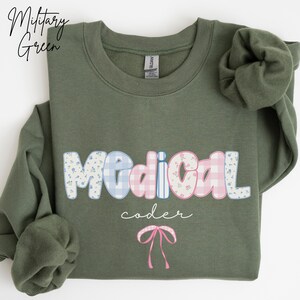 May include: Military green sweatshirt with the words "Medical coder" in a playful font. The word "Medical" is in a bubble letter style with floral and gingham patterns. A pink bow graphic is at the bottom.