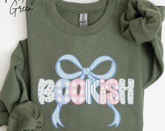 Puede incluir: Sudadera verde oliva con la palabra "BOOKISH" en una fuente caprichosa, adornada con un lazo azul. Las letras están decoradas con diseños florales, cuadros y rayas. El texto "Military Green" está en la esquina superior izquierda.
