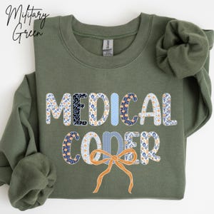 May include: Military green sweatshirt with the words "MEDICAL CODER" in a mix of patterned fonts. The letters are decorated with floral, leopard, and striped patterns. An orange bow is at the bottom.
