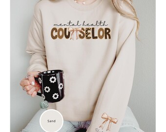 May include: A sand-colored sweatshirt with the text "mental health counselor" in brown and black letters with a brown bow. The sweatshirt has a brown bow on the sleeve with the text "Victoria" underneath.