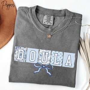 May include: A gray Comfort Colors t-shirt with the word "DOULA" in white block letters, each letter set on a light blue background with a floral pattern. A blue bow graphic is below the word. A silver necklace with a small pendant is on the shirt.