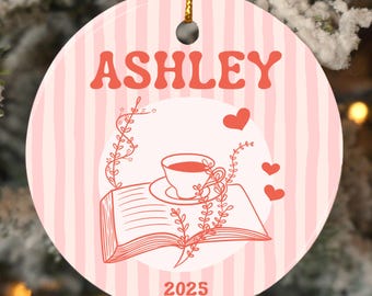 May include: Round ceramic ornament with a pink and white striped background. The ornament features the name "ASHLEY" in orange, along with a drawing of a teacup, book, hearts, and the year "2025". A gold string is attached for hanging.