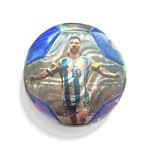 May include: A soccer ball featuring a metallic, holographic finish with blue panels. The central panel displays a photographic print of a soccer player wearing a light blue and white striped jersey with the number 10, arms outstretched.