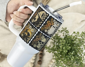 May include: White tumbler with a handle and a straw. The tumbler features four square designs with a witch, a fox, a ghost, and a skull, all set against a floral and checkered background. The design is in shades of black, white, and orange.