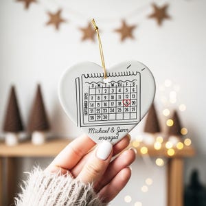 May include: A white heart-shaped ceramic ornament with a calendar graphic for September 2025, highlighting the 19th. The text "Michael & Jenn engaged" is below the calendar. A gold string is attached for hanging.