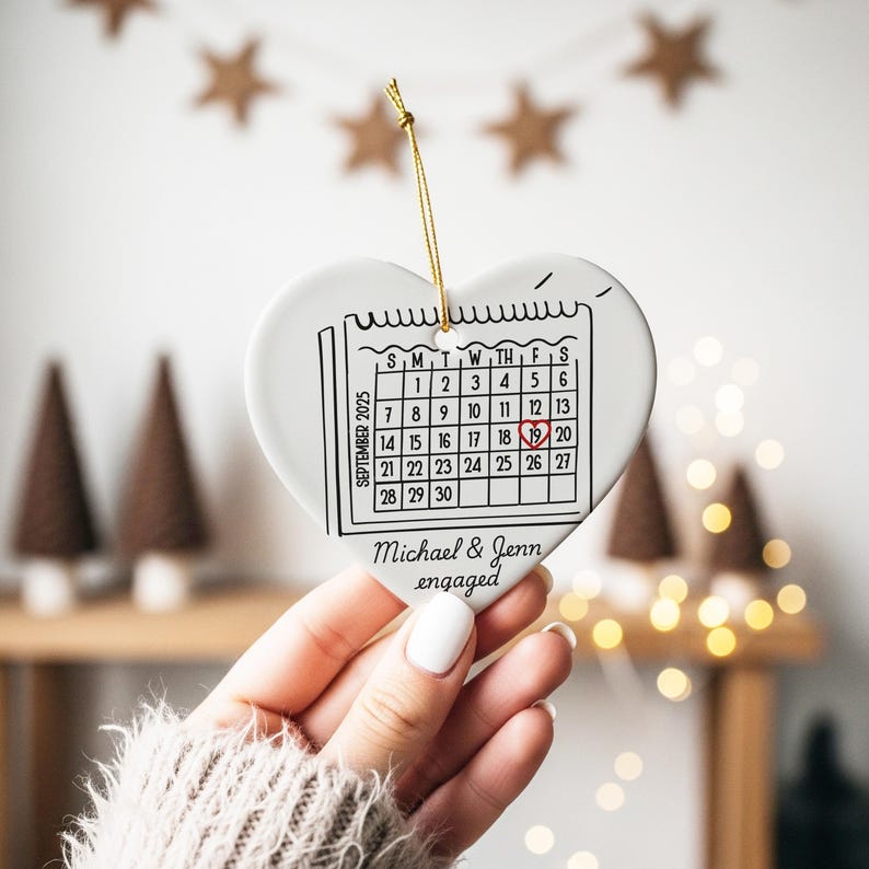 May include: A white heart-shaped ceramic ornament with a calendar graphic for September 2025, highlighting the 19th. The text "Michael & Jenn engaged" is below the calendar. A gold string is attached for hanging.