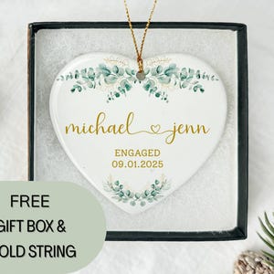 May include: A white heart-shaped ceramic ornament with gold lettering that reads "michael & jenn" and "ENGAGED 09.01.2025". The ornament is decorated with green and gold floral accents and comes in a black gift box.