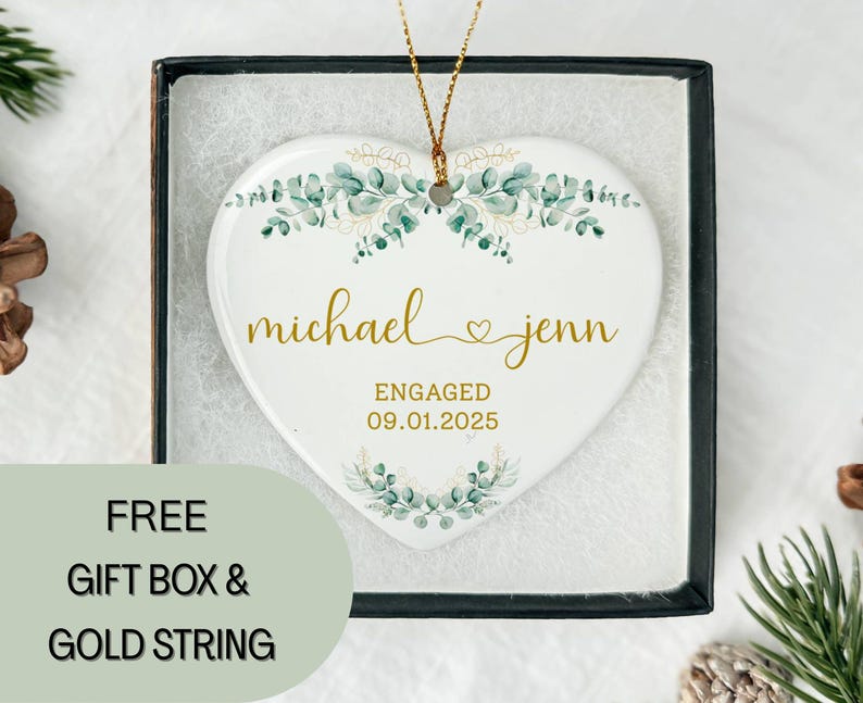 May include: A white heart-shaped ceramic ornament with gold lettering that reads "michael & jenn" and "ENGAGED 09.01.2025". The ornament is decorated with green and gold floral accents and comes in a black gift box.