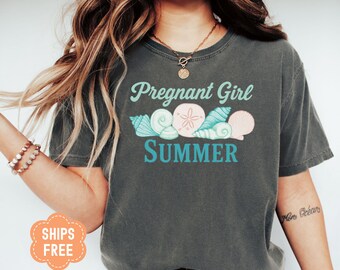 May include: Dark gray t-shirt with the words "Pregnant Girl Summer" in teal and white script. The design includes illustrations of seashells and a sand dollar. The shirt is a relaxed fit.