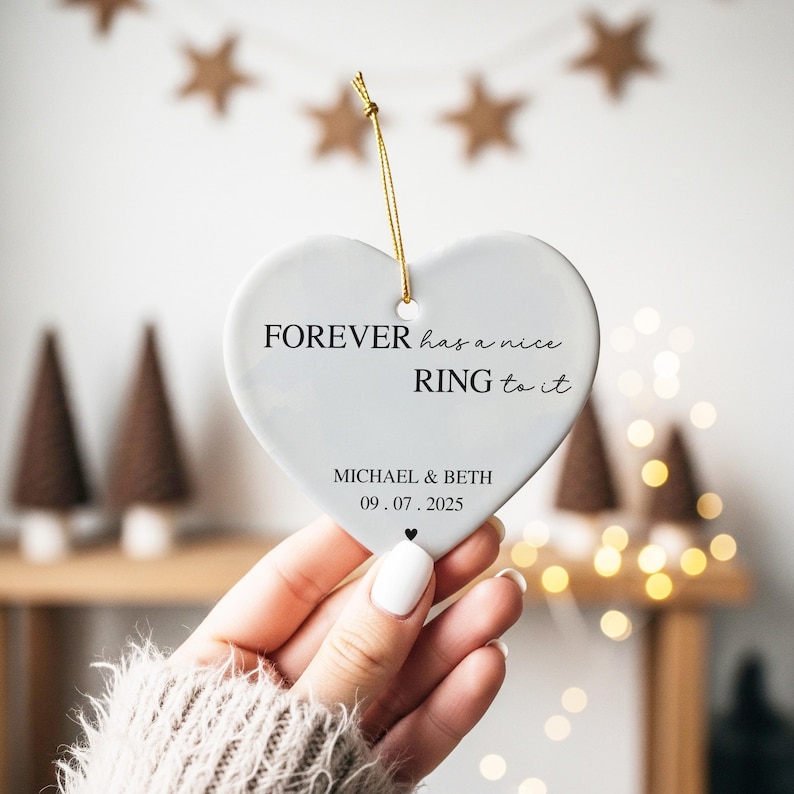 May include: A heart-shaped, white ceramic ornament with the text "FOREVER has a nice RING to it." and the names "MICHAEL & BETH" with the date "09.07.2025." A gold string is attached for hanging. Decorative item.