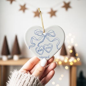 May include: A white heart-shaped ornament with blue ink drawings of a bow, two hearts, and the word "engaged." One heart has the initials "M&A," and the other has the year "2025." A gold string is attached for hanging.