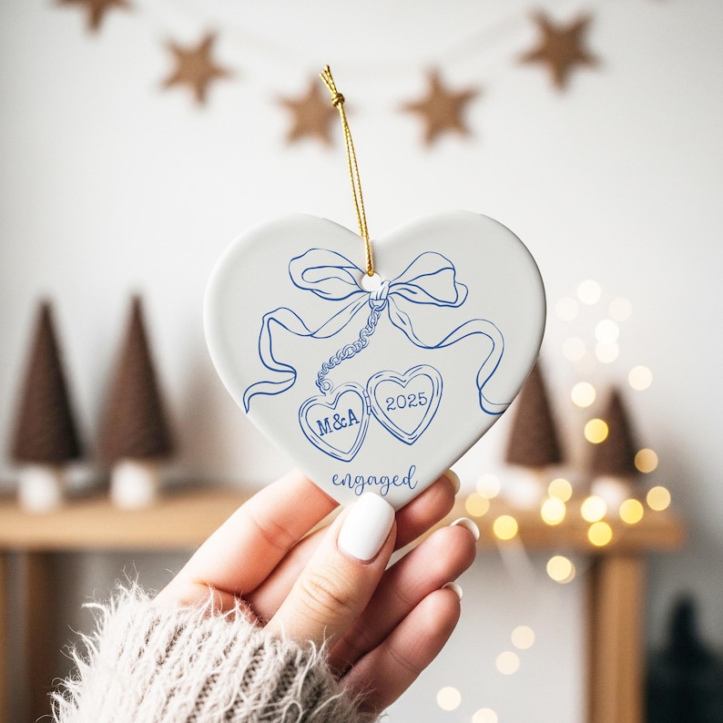 May include: A white heart-shaped ornament with blue ink drawings of a bow, two hearts, and the word "engaged." One heart has the initials "M&A," and the other has the year "2025." A gold string is attached for hanging.