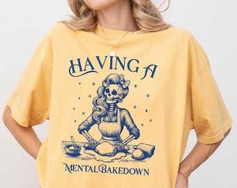 May include: Mustard yellow t-shirt with a navy blue graphic. The graphic features a skeleton woman baking with the text "HAVING A MENTAL BAKEDOWN". The design includes stars and a rolling pin.