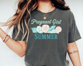 May include: Dark gray t-shirt with the text "Pregnant Girl Summer" in teal and white. The design includes illustrations of seashells and a sand dollar. The shirt is a casual, comfortable style.