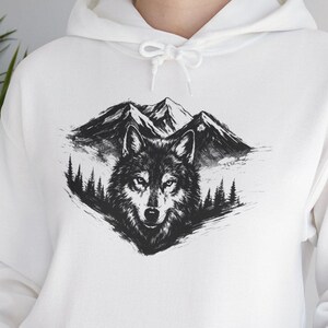 Puede incluir: Sudadera con capucha blanca con un diseño gráfico en blanco y negro de un lobo, montañas y árboles. El lobo es el motivo central. La sudadera tiene un bolsillo delantero y una capucha con cordón.