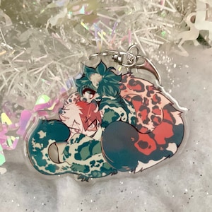 Peut inclure: Un porte-clés en acrylique transparent avec une illustration colorée de style anime. Le design comprend trois personnages aux cheveux turquoise, rouges et blancs, et une queue avec un motif léopard. Le porte-clés a un fermoir argenté.