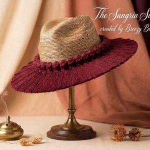Puede incluir: Un sombrero fedora de paja con un borde de flecos color burdeos oscuro y una fila de nudos de cuerda a juego alrededor de la base de la copa. El texto The Sangria Sunset created by Breezy Bohemian es visible en la esquina superior derecha.
