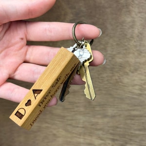 May include: A wooden keychain with a heart and the letters "D & A" engraved on it. The keychain also has the text "Drive Safe I need you here with me".