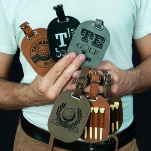 May include: Four brown leather luggage tags with golf-themed designs. One tag features a golf ball and the text "Thomas Est. 2018". Another tag has a golf club and the text "T. Thomas". A third tag has the text "TE Golf" and a fourth tag has a golf ball and the text "Thomas" with a laurel wreath.