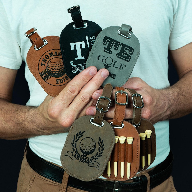 May include: Four brown leather luggage tags with golf-themed designs. One tag features a golf ball and the text "Thomas Est. 2018". Another tag has a golf club and the text "T. Thomas". A third tag has the text "TE Golf" and a fourth tag has a golf ball and the text "Thomas" with a laurel wreath.