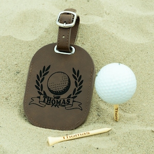 May include: Brown leather luggage tag with a golf ball and wreath design. The tag is personalized with the name "Thomas".