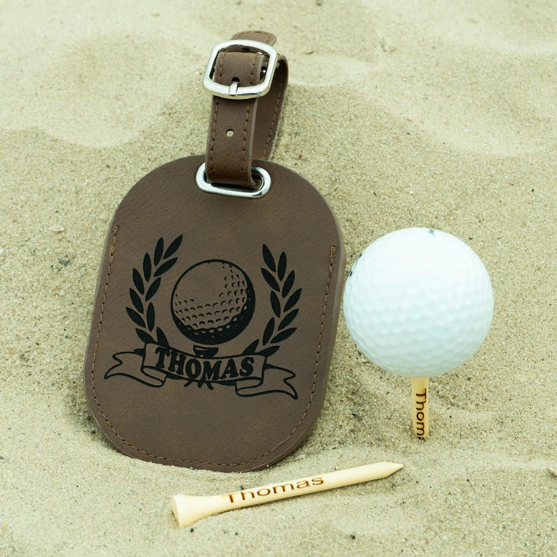 May include: Brown leather luggage tag with a golf ball and wreath design. The tag is personalized with the name "Thomas".