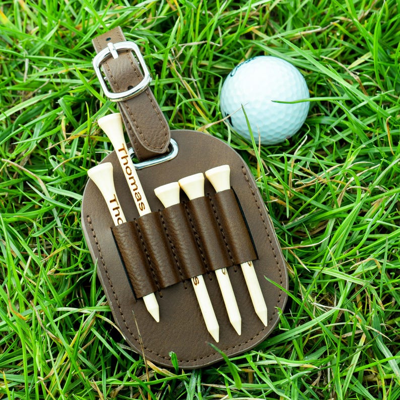 May include: Brown leather golf tee holder with the name "Thomas" engraved on it. The holder has five slots for golf tees and is attached to a golf bag.
