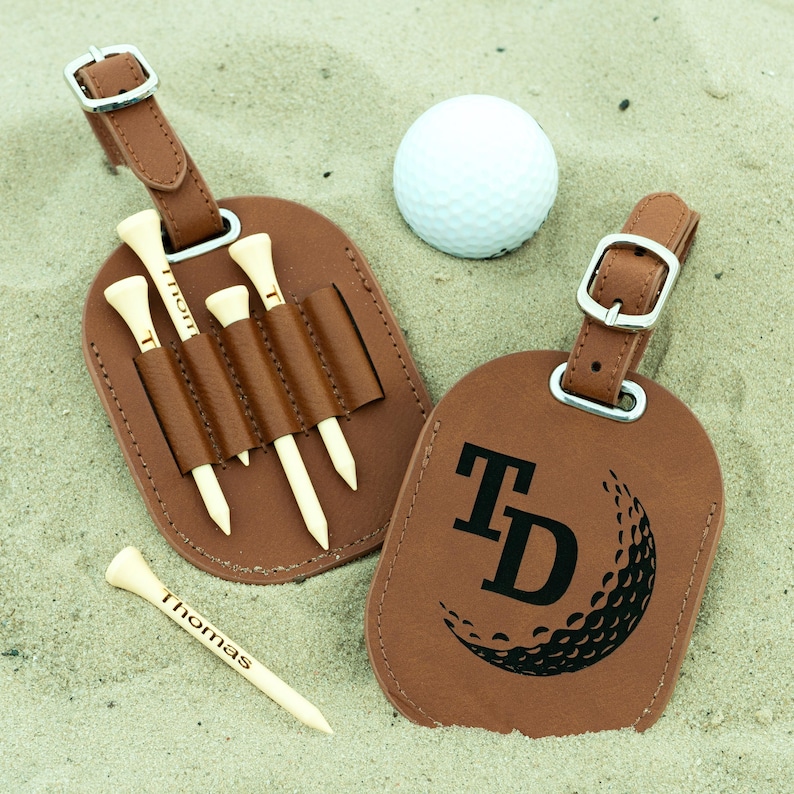 May include: Two brown leather luggage tags. One tag has a golf ball design with the initials "TD" and the other tag has four slots for golf tees. The tag with the golf tees has the name "Thomas" engraved on a tee.