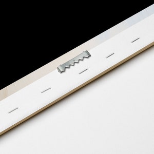 May include: Close-up of the back of a canvas print with a metal hanging hook and staples. The canvas is white and the wood frame is light brown.