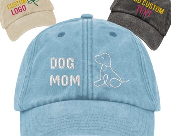 May include: A light blue denim baseball cap with the text "DOG MOM" embroidered in white on the front. A white outline of a dog is embroidered to the right of the text.