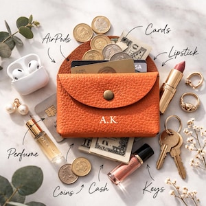 Puede incluir: Una cartera de cuero naranja con las iniciales "A.K." grabadas en la parte delantera, abierta para revelar efectivo y tarjetas. La cartera está rodeada de accesorios, incluyendo AirPods, monedas, lápiz labial, anillos, perfume, brillo labial, llaves y aretes.