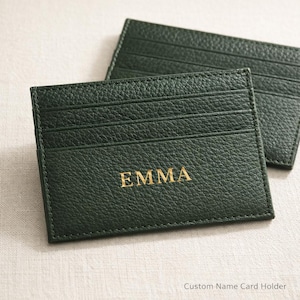 Puede incluir: Dos tarjeteros de cuero verde oscuro. El tarjetero frontal tiene el nombre "EMMA" en letras doradas. El tarjetero tiene varias ranuras para tarjetas. El fondo es de color beige claro.
