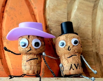 Op de afbeelding: Twee wijnkurken verkleed als bruid en bruidegom. De bruid draagt een paarse hoed en de bruidegom draagt een zwarte hoge hoed. Ze zitten op een houten oppervlak met een pompoen op de achtergrond.