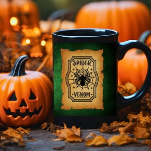May include: Black coffee mug with a green background and a vintage label that says "Spider Venom" with a black spider and web graphic. The mug is sitting on a table with fall leaves and pumpkins.