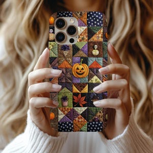 May include: A patterned phone case with a patchwork design. The case features a jack-o'-lantern, pumpkins, and autumn leaves. The case is held in front of a person wearing a white sweater. The case has a Halloween theme.