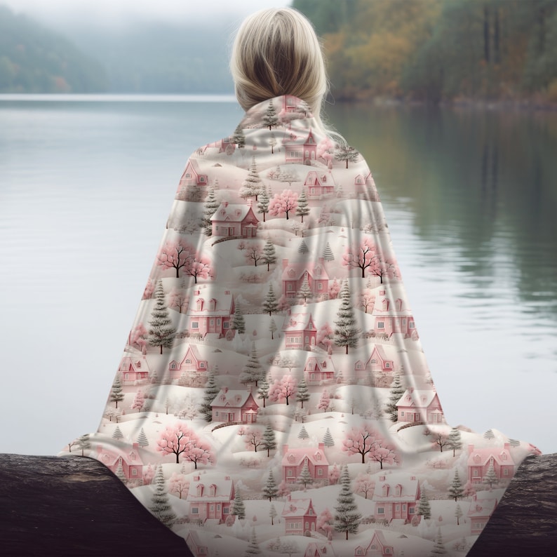 May include: A white blanket with a pink and green winter village pattern. The blanket is draped over a log and has a snowy background.