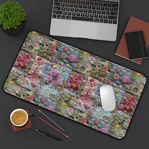Pode incluir: Tapete de rato bordado com design floral e borda preta. O padrão colorido apresenta flores e folhas rosa, azuis e verdes. Um rato de computador branco está sobre o tapete, ao lado de um laptop, um caderno e uma xícara de café.