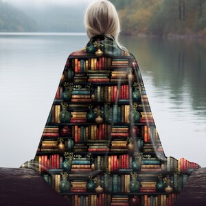 May include: A soft, cozy blanket featuring a repeating pattern of bookshelves filled with colorful books and decorative vases. The blanket is draped over a wooden log, with a lake and trees in the background.
