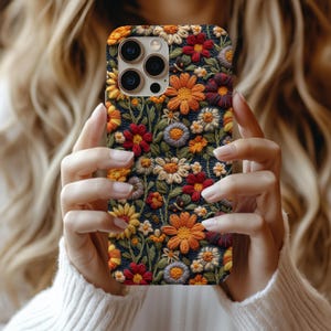 Puede incluir: Una funda de teléfono con un diseño floral bordado. La funda presenta un fondo oscuro con una variedad de flores coloridas en tonos de amarillo, naranja, rojo y blanco. La funda se sostiene frente a una persona.