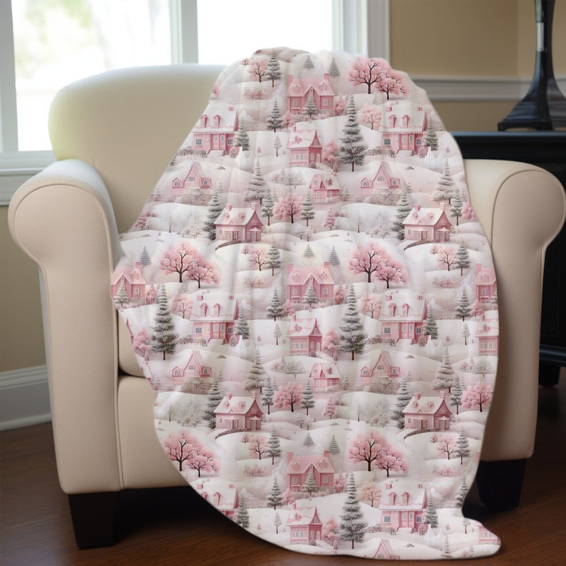 May include: A pink and white throw blanket with a repeating pattern of houses, trees, and snow. The houses are pink with white trim and the trees are green with white snow on the branches.