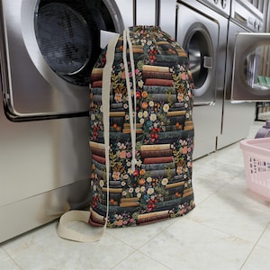 Puede incluir: Una bolsa de lavandería con cordón con un estampado floral y de libros. La bolsa presenta un fondo oscuro con pilas de libros y flores coloridas. Tiene una correa beige y está cerca de una lavadora.
