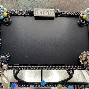 May include: Black metal picture frame with a chain border and a silver plaque that says "Laugh". The frame is decorated with blue, white, and silver rhinestones, pearls, and skull charms.