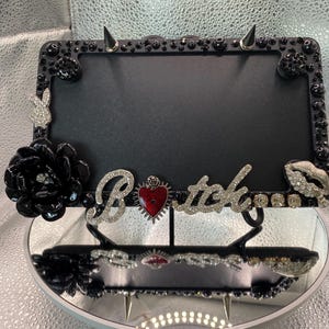 May include: A rectangular black license plate frame decorated with black beads, silver spikes, a rhinestone rabbit silhouette, a large black flower, and a silver rhinestone script spelling the word Bitch with a red heart-shaped charm in the center.