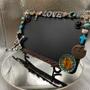 May include: Black picture frame with decorative embellishments. The frame is adorned with charms, including crosses, the word "LOVE", and turquoise beads. A bottle cap with a religious image is also attached.