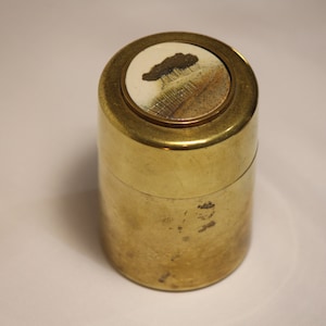 May include: A cylindrical brass container with a lid. The lid has a circular inset with a painted landscape scene. The container has a smooth, polished surface and a slightly aged appearance.