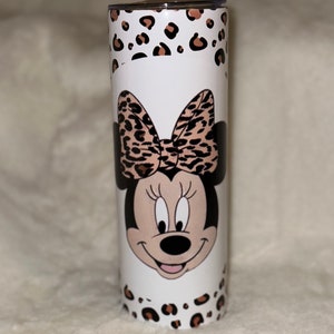 May include: A white tumbler featuring Minnie Mouse with a leopard print bow and accents. The tumbler has a stainless steel appearance with a lid. The design includes a leopard print pattern around the top and bottom.